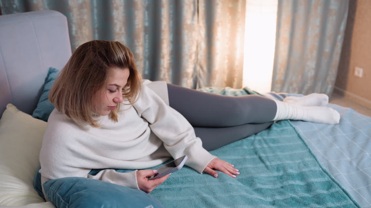 Woman relaxing on bed in cozy sweater and leggings holding smartphone, looking at screen with calm expression, resting on soft pillows in warm home interior with curtain and lamp providing ambient light