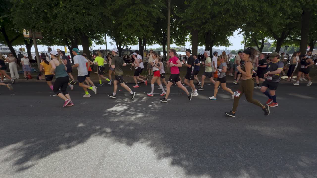 Runners participate in Stockholm Marathon on sunny day, surrounded by lush green trees