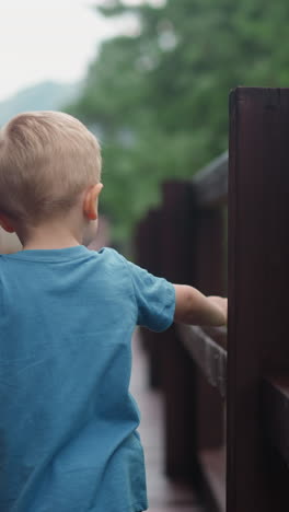 curious little boy touches wet wooden railing made of large beams on deck at cottage house after warm spring rain side view slow motion
