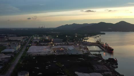 aerial tracking shot capturing bulk carrier container ships docked at lumut port industrial park on sungai manjung river at dusk, kampung acheh, perak, malaysia, mountain landscape in the background