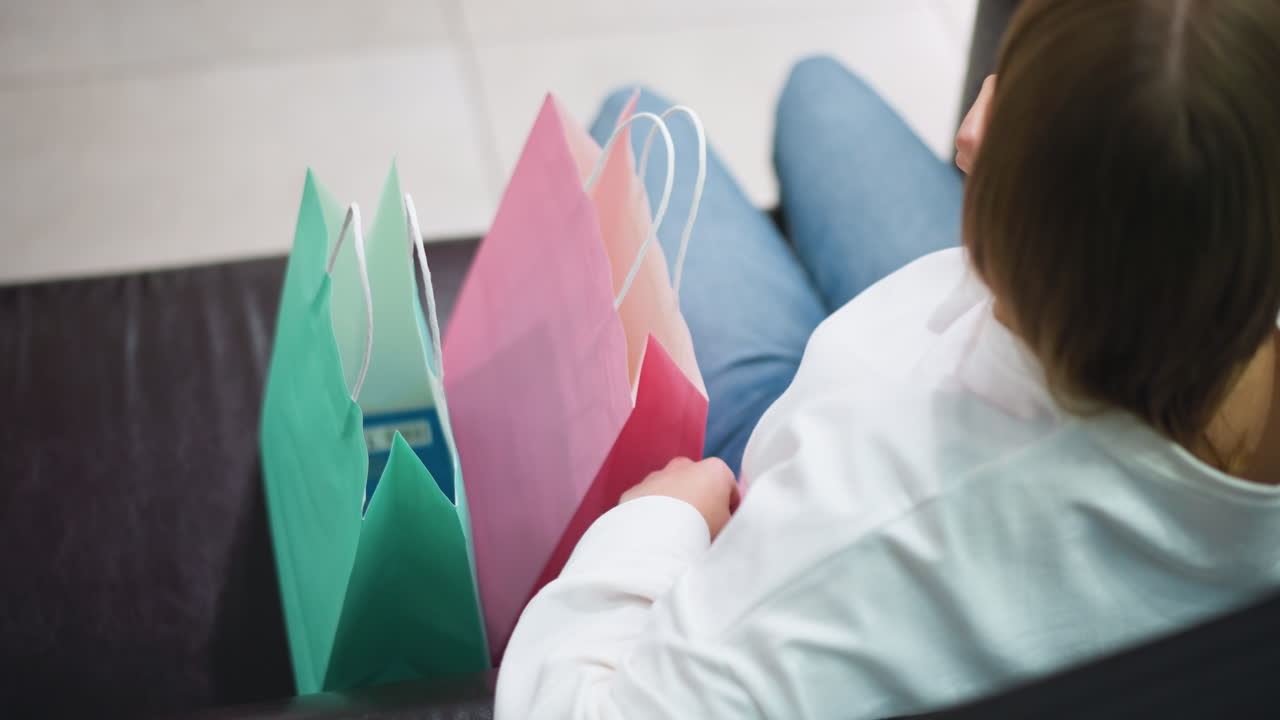 vista trasera de la dama sentada en el sofá sosteniendo bolsas de compras de diferentes colores, ajusta su cabello, bolsas de rosa, verde y azul se colocan en su regazo mientras se relaja