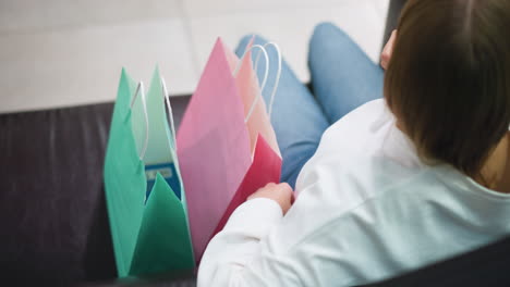 rear view of lady seated on couch holding shopping bags in different colors, adjust her hair, bags in pink, green, and blue are placed on her lap while she relaxes