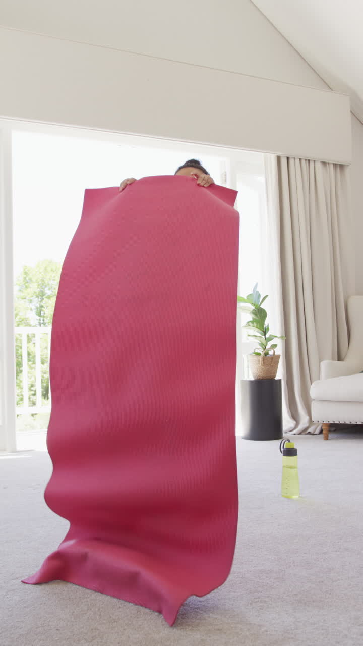 Vertical video of happy asian woman placing yoga mat in bedroom, in slow motion