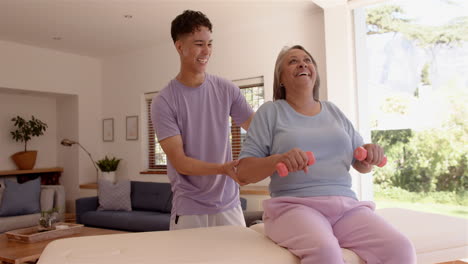 assisting senior woman with arm exercises, male physiotherapist smiling in living room