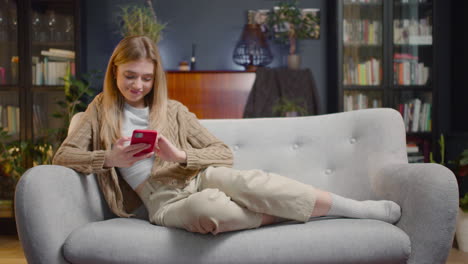 Happy-Young-Woman-Using-Smartphone-While-Lying-On-A-Couch-At-Home