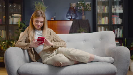 Mujer-Joven-Feliz-Usando-Un-Teléfono-Inteligente-Mientras-Está-Acostado-En-Un-Sofá-En-Casa