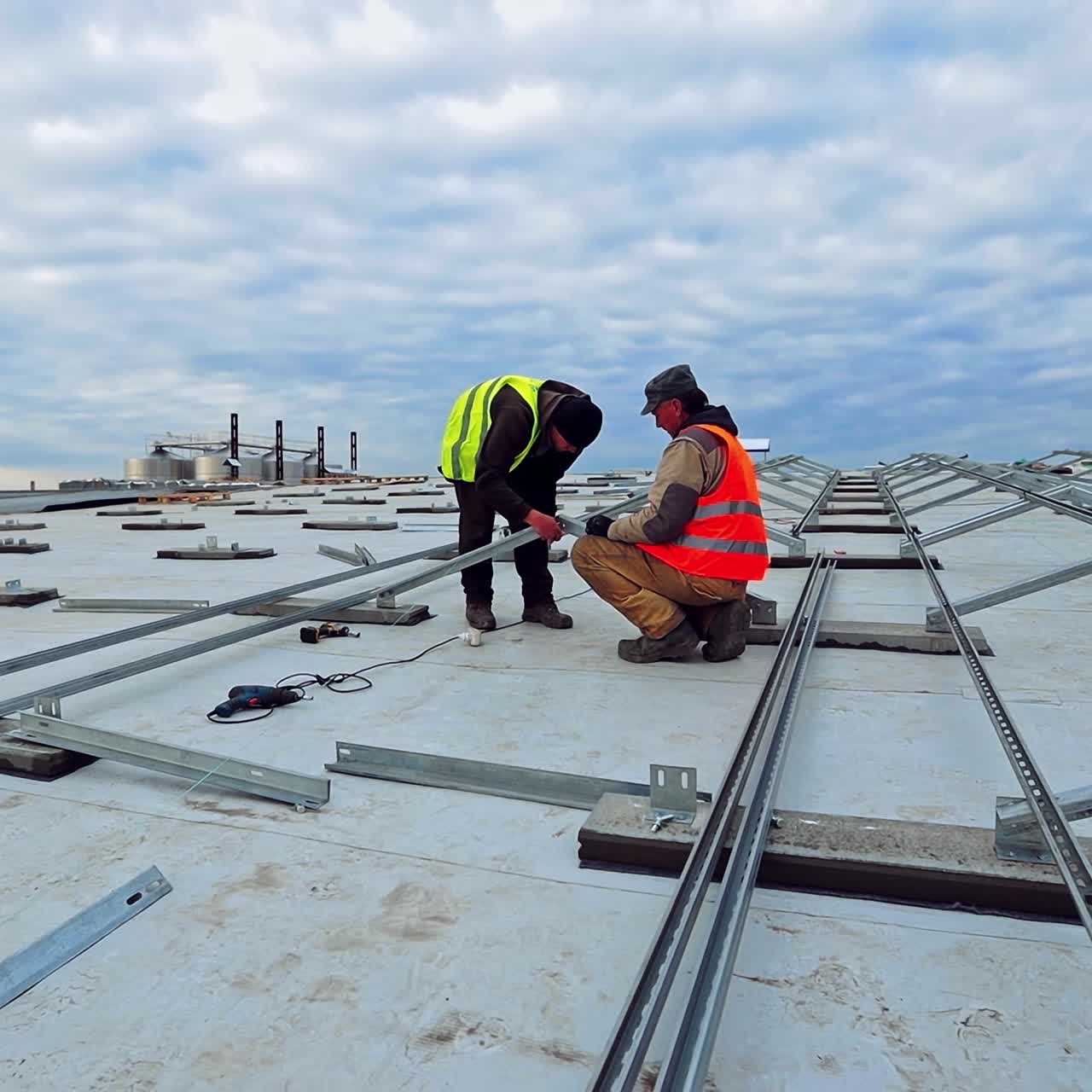 New construction of solar farm on the top of building. Two workers in colorful waistcoats attaching metal base on a flat roof.