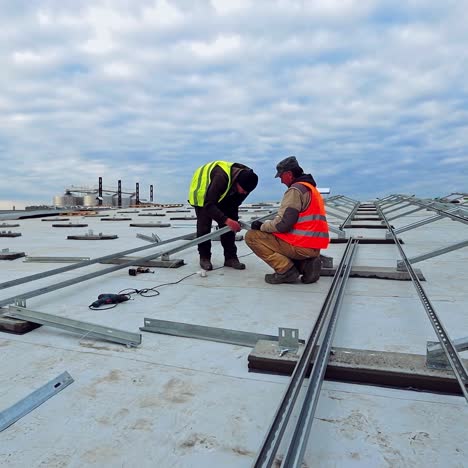 new construction of solar farm on the top of building. two workers in colorful waistcoats attaching metal base on a flat roof.