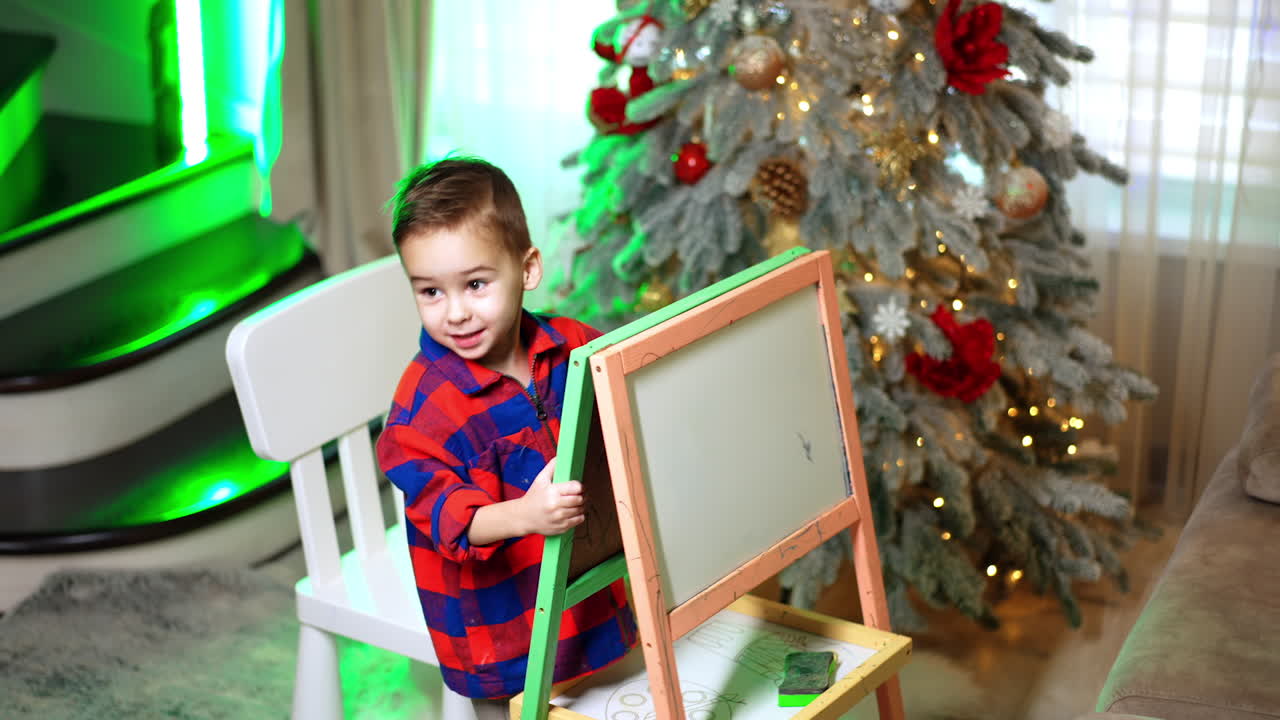 Child standing near Christmas tree with board. Little boy in red plaid shirt standing near Christmas tree and drawing board in cozy living room