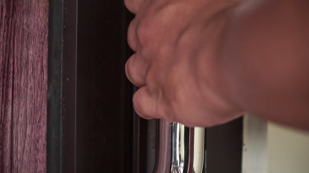 Human hand turning and pulling a shiny door handle to close a wooden door. Detailed close-up showing texture of skin and door material