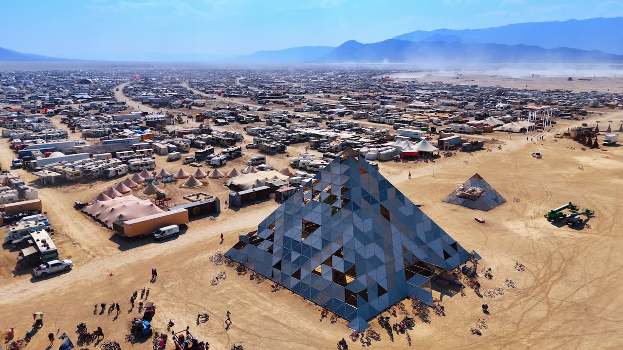 Reflective pyramid beside desert camps. A mirrored pyramid stands among tents and vehicles under bright light on the open playa