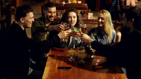 cheerful young men and women are celebrating important event in nice bar. they are clanging glasses, toasting and drinking. friends having fun at weekend concept.