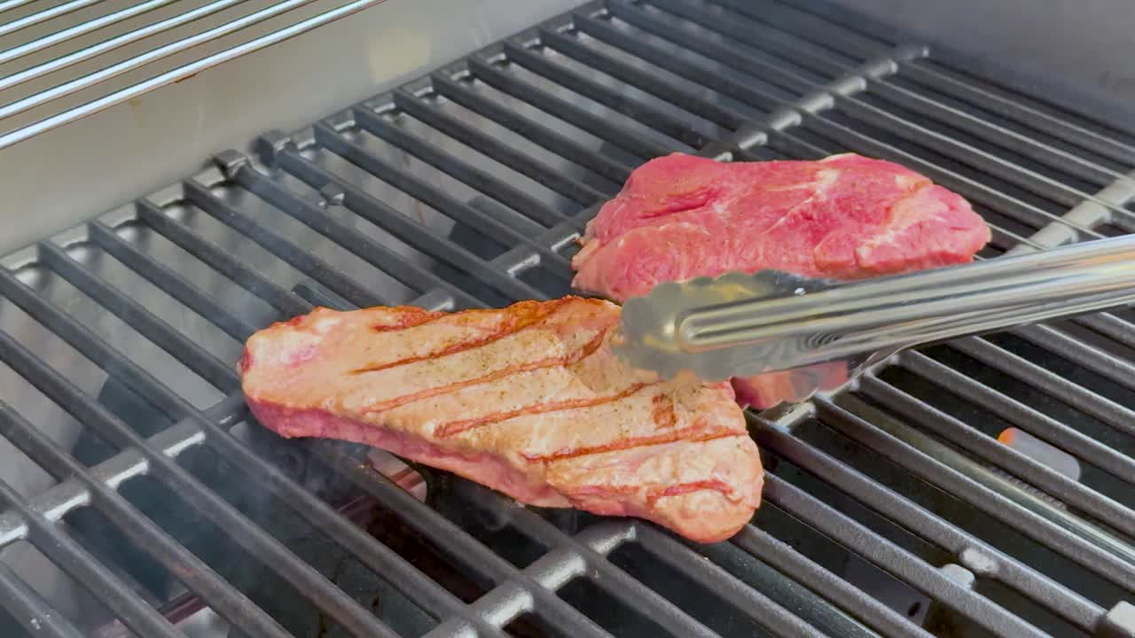 Two raw beef steaks are turned with metal tongs on a gas grill, developing grill marks as smoke rises in bright, natural lighting