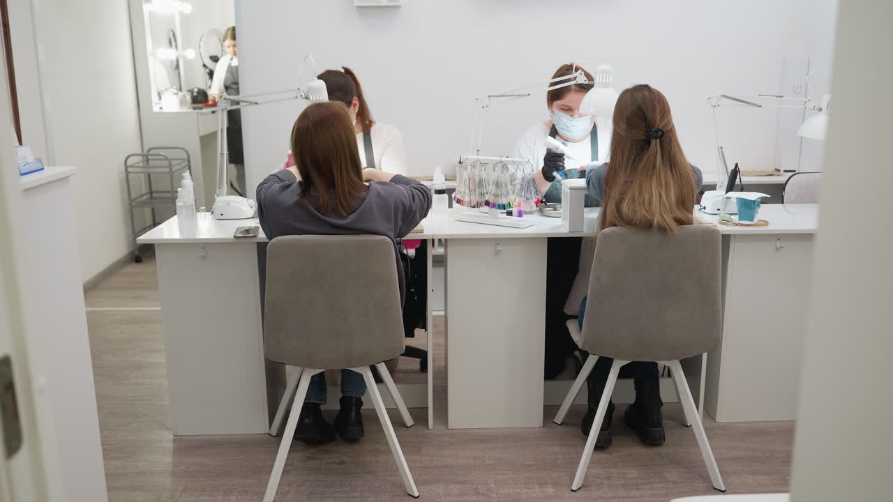 Two women with blond hair seated on peach chairs with white legs, wearing jeans and black shoes, face nail technicians while receiving manicure services in bright modern salon with nail tools