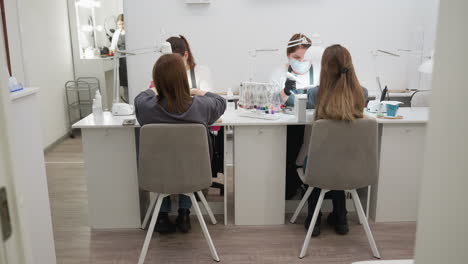 two women with blond hair seated on peach chairs with white legs, wearing jeans and black shoes, face nail technicians while receiving manicure services in bright modern salon with nail tools