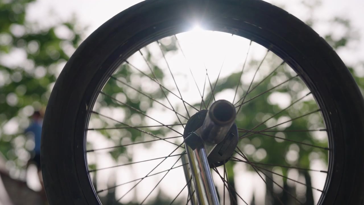 Detail předního kola BMX s malou hloubkou ostrosti, zatímco v pozadí je rozmazané dítě jedoucí na koloběžce ve skateparku. Scéna z městského životního stylu.