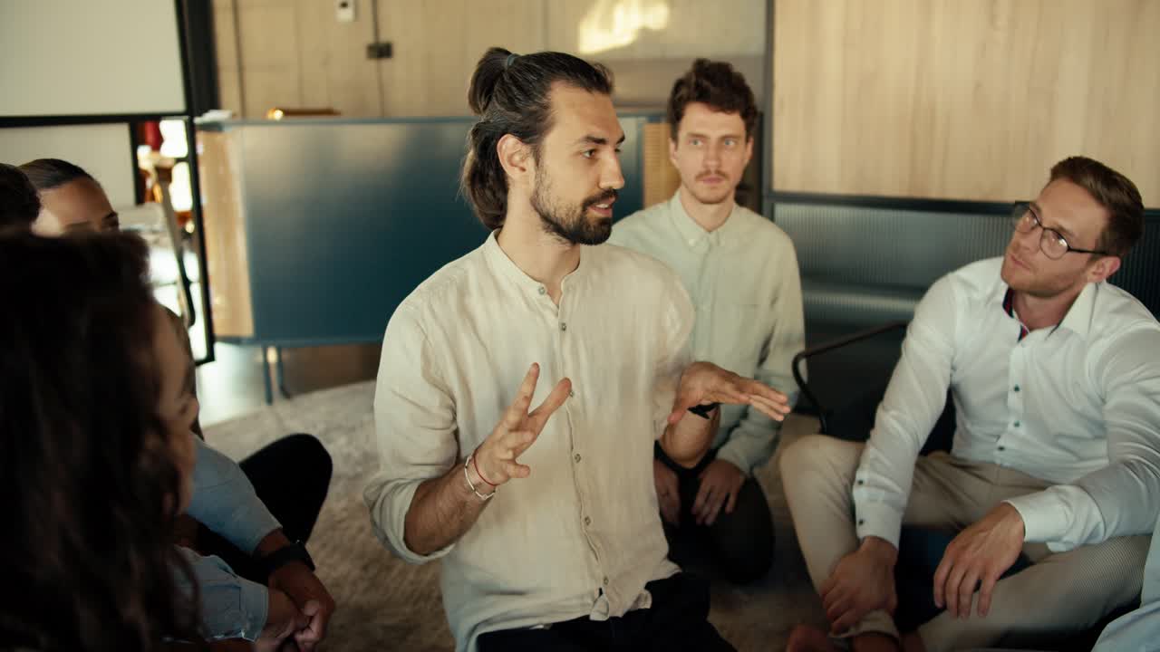A man in a shirt talks about his problem at a group therapy in the office. The rest of the participants in therapy listen to the man and stretch their hands to him in order to support him