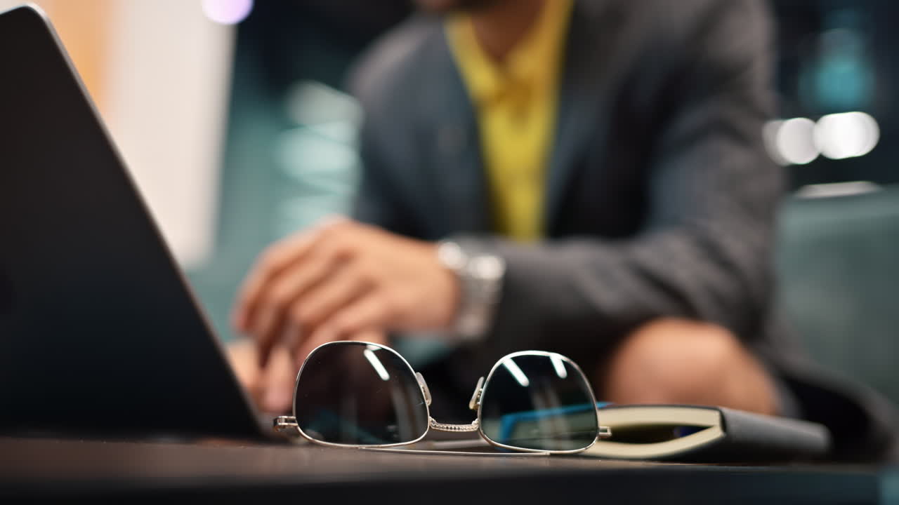 Close up of a pair of sunglasses standing on a table with a man working on a laptop on the background