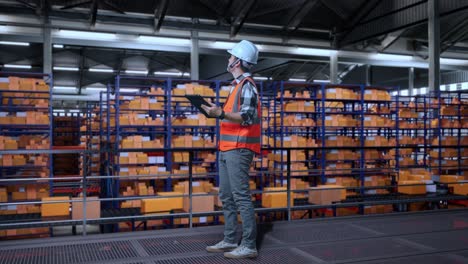 vista lateral completa del cuerpo de un ingeniero masculino asiático con casco de seguridad trabajando en una tableta mientras estaba de pie en el almacén con estantes llenos de productos de entrega