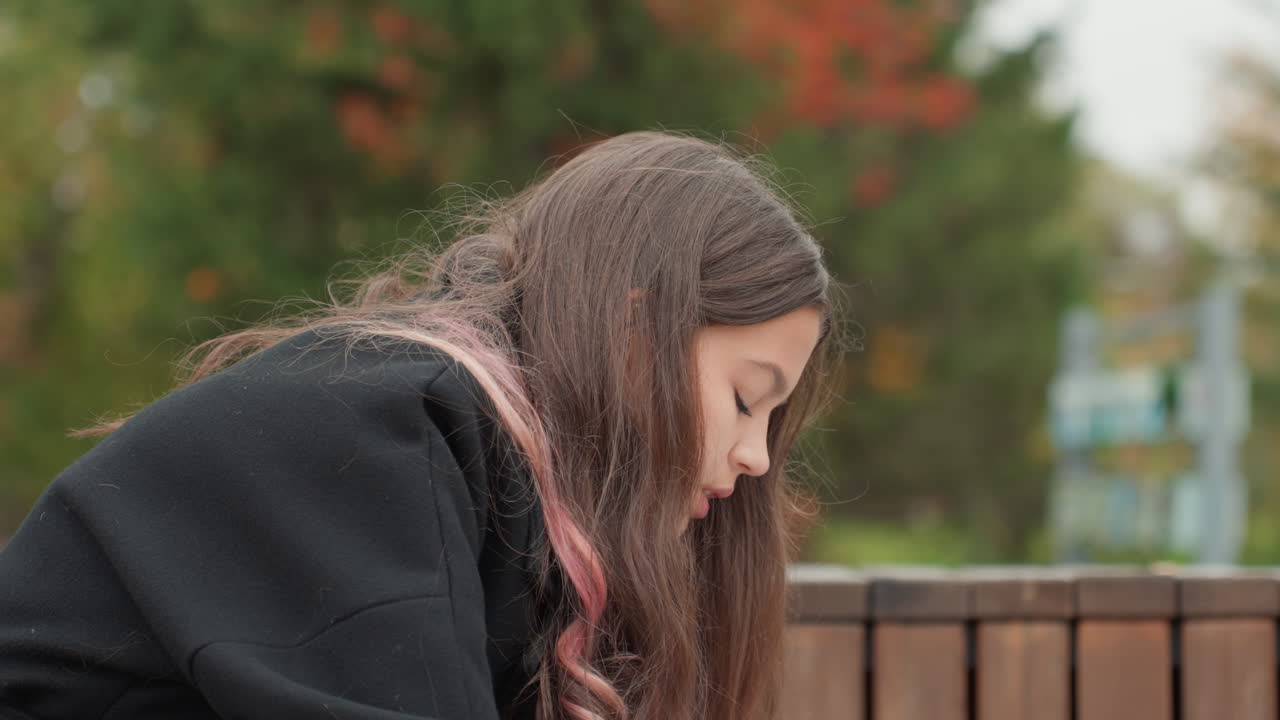 Estudante caucasiana adolescente lendo em um banco, expressão concentrada, cabelo castanho comprido com mechas rosas, casaco preto, folhagem de outono com plano de fundo desfocado, ambiente de campus em parque, estudando em um caderno, silencioso