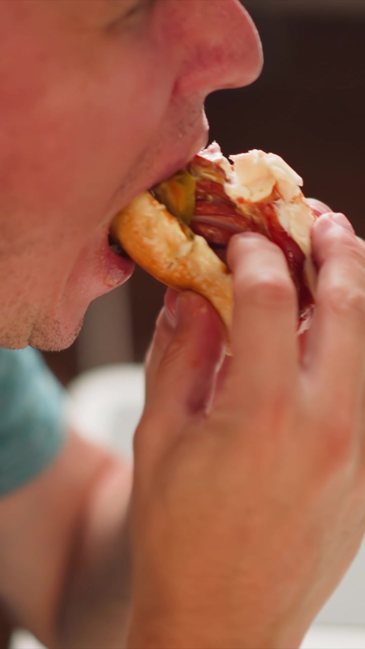 Close up of man eating hot dog with mustard and ketchup. Hand holding food while taking bites showing texture detail on bun toppings. Indoor setting with soft lighting and shallow depth