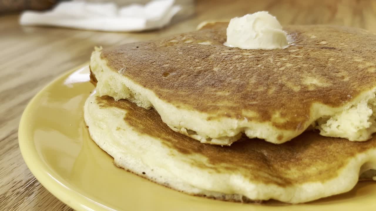 A mouthwatering close-up of golden syrup being poured over fluffy pancakes with melting butter, captured in a cozy cafe or diner setting.