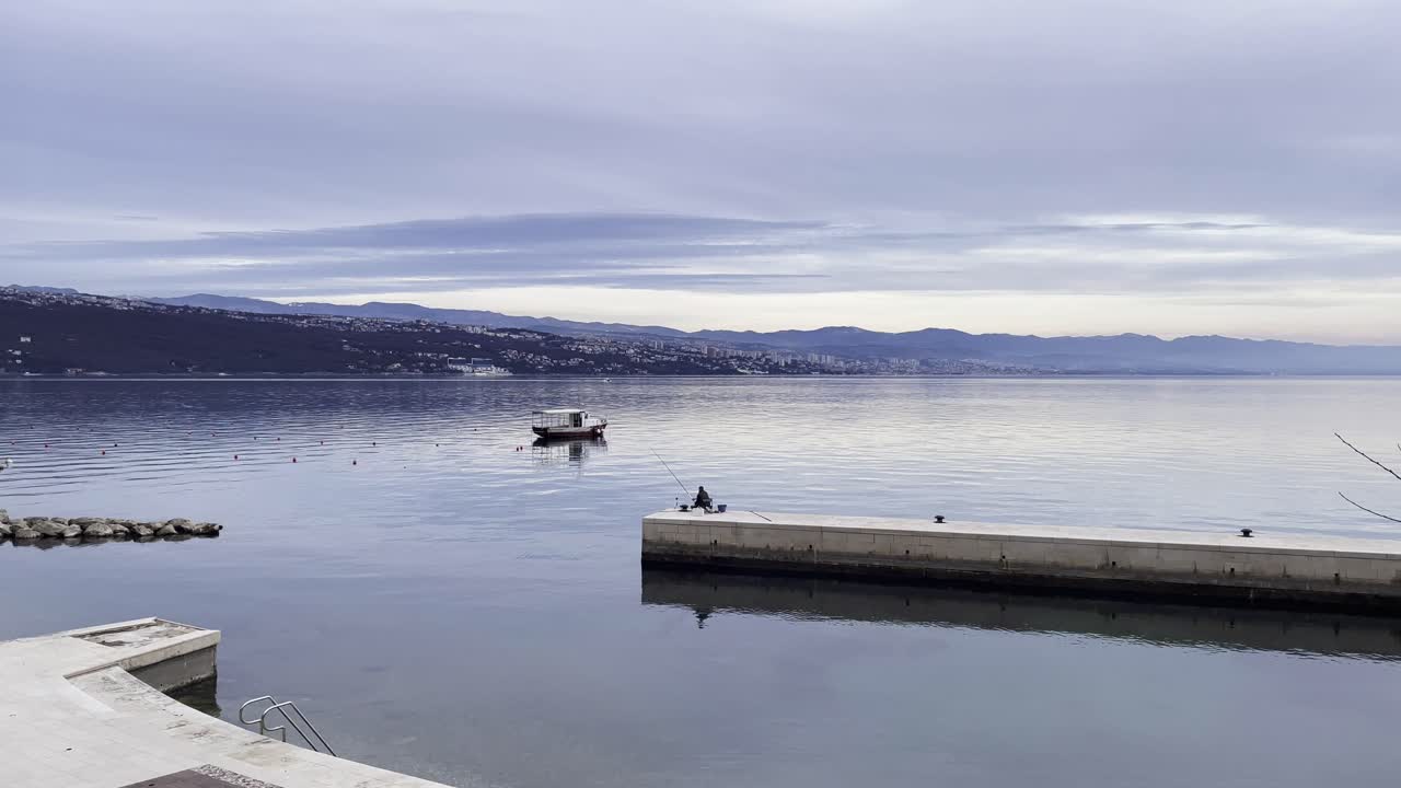 Fisherman casting a line on peer Boat parked on Sea shore with town in distance wide, Opatija, Croatia