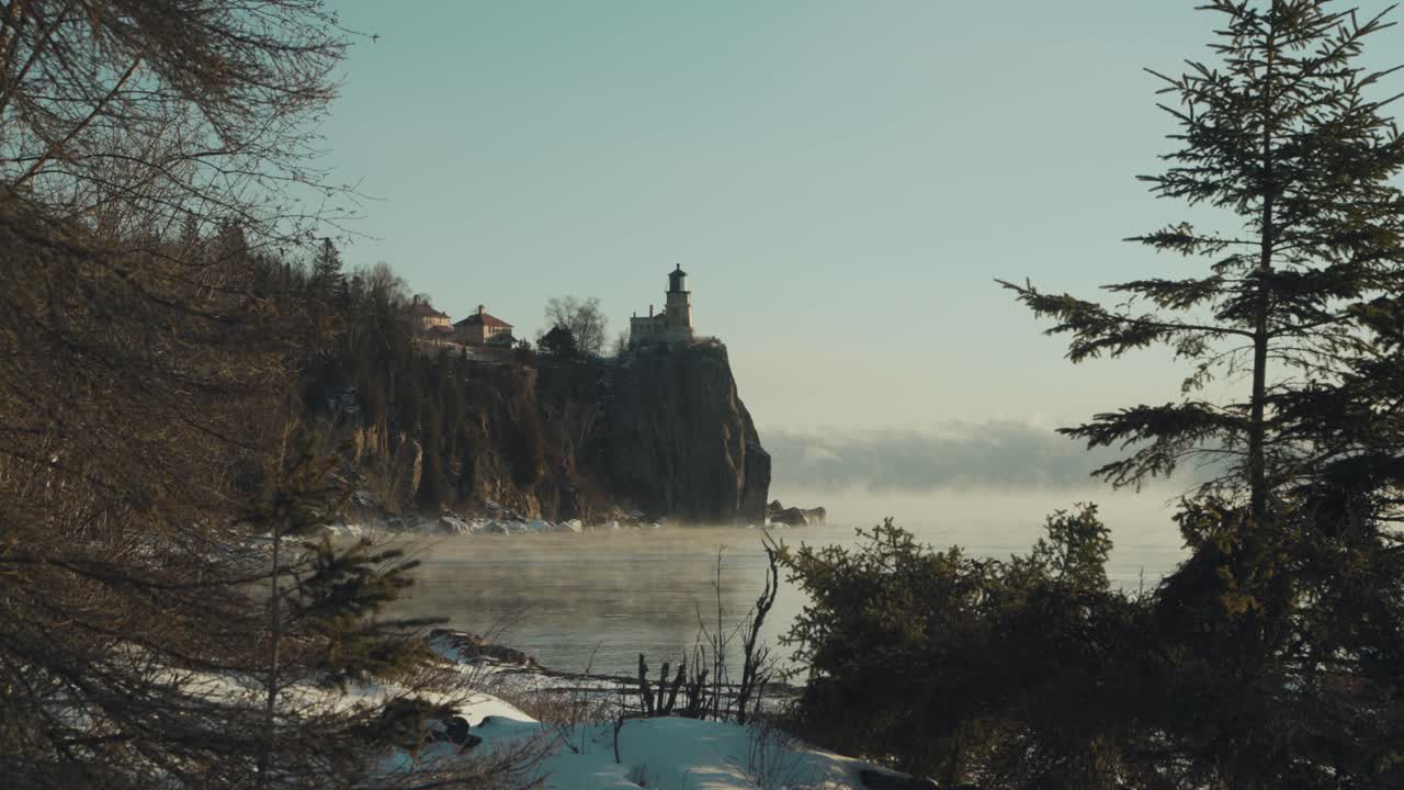 Ampia inquadratura cinematografica dello storico faro di Split Rock su un'alta scogliera, con una densa nebbia marina che sale dal Lago Superiore in una gelida mattina del Minnesota.