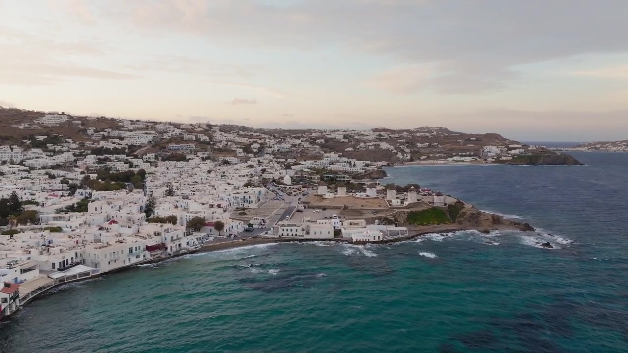 Mulini a vento dell'isola di Mykonos e Piccola Venezia con acqua turchese ed edifici bianchi sotto un cielo tenue, panoramica aerea, ampia inquadratura di ambientazione costiera