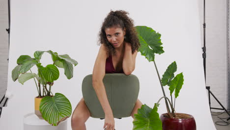 dark curly haired and tanned model posing on a chair with a red bikini suit, surrounded by plants, shot in a photo studio