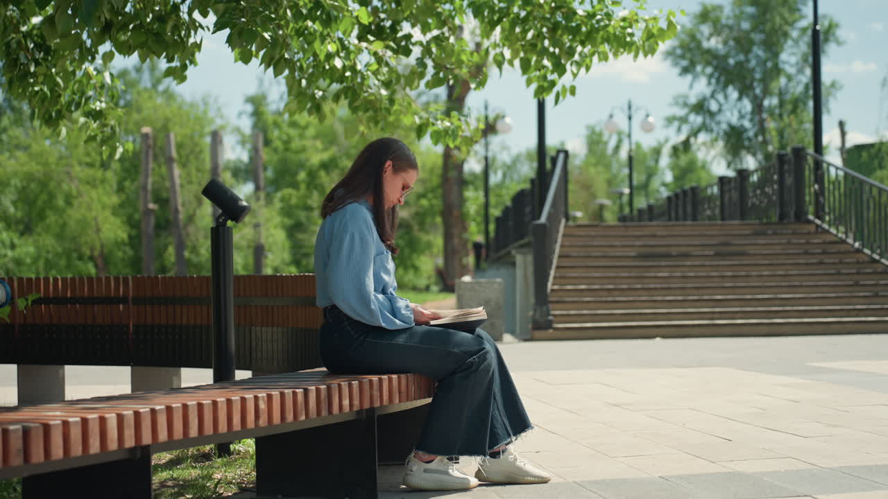 계단 근처 벤치에서 책을 읽고 있는 젊은 백인 여성, 도시 공원에서 가이드북을 보며 즐거워하는 사려 깊은 여행객 또는 학생, 잎이 무성한 덮개와 콘크리트 계단, 부드러운 한낮의 햇살 아래 편안한 자세