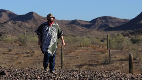 en el desierto de sonora, un hombre desesperado vestido con un poncho, un pañuelo rojo y un sombrero de vaquero camina por un paisaje accidentado lleno de rocas y cactus saguaro.