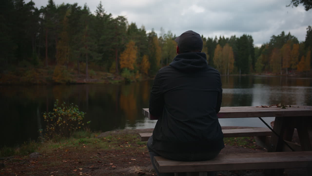 A lonely man sits on a bench in the woods looking out to a lake and enjoying the view