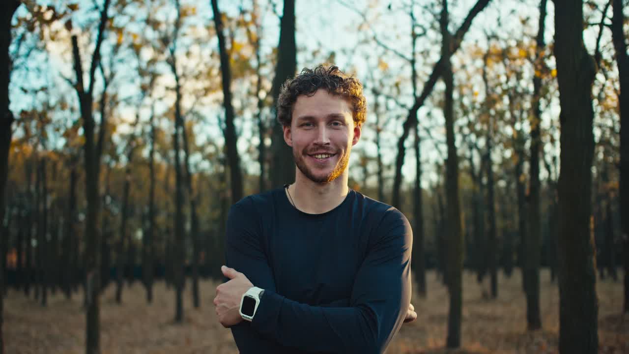 Portrait of a happy and confident man with curly hair a brunette with a beard who crossed his arms on his chest and stands confidently while jogging in the autumn forest in the morning