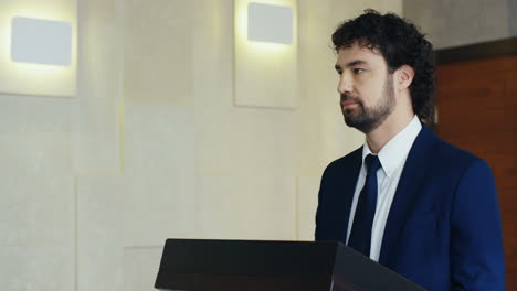 Close-up-view-of-caucasian-businessman-speaker-on-a-podium-wearing-formal-clothes-and-talking-in-a-conference-room,-then-he-turns-and-smiles-at-camera