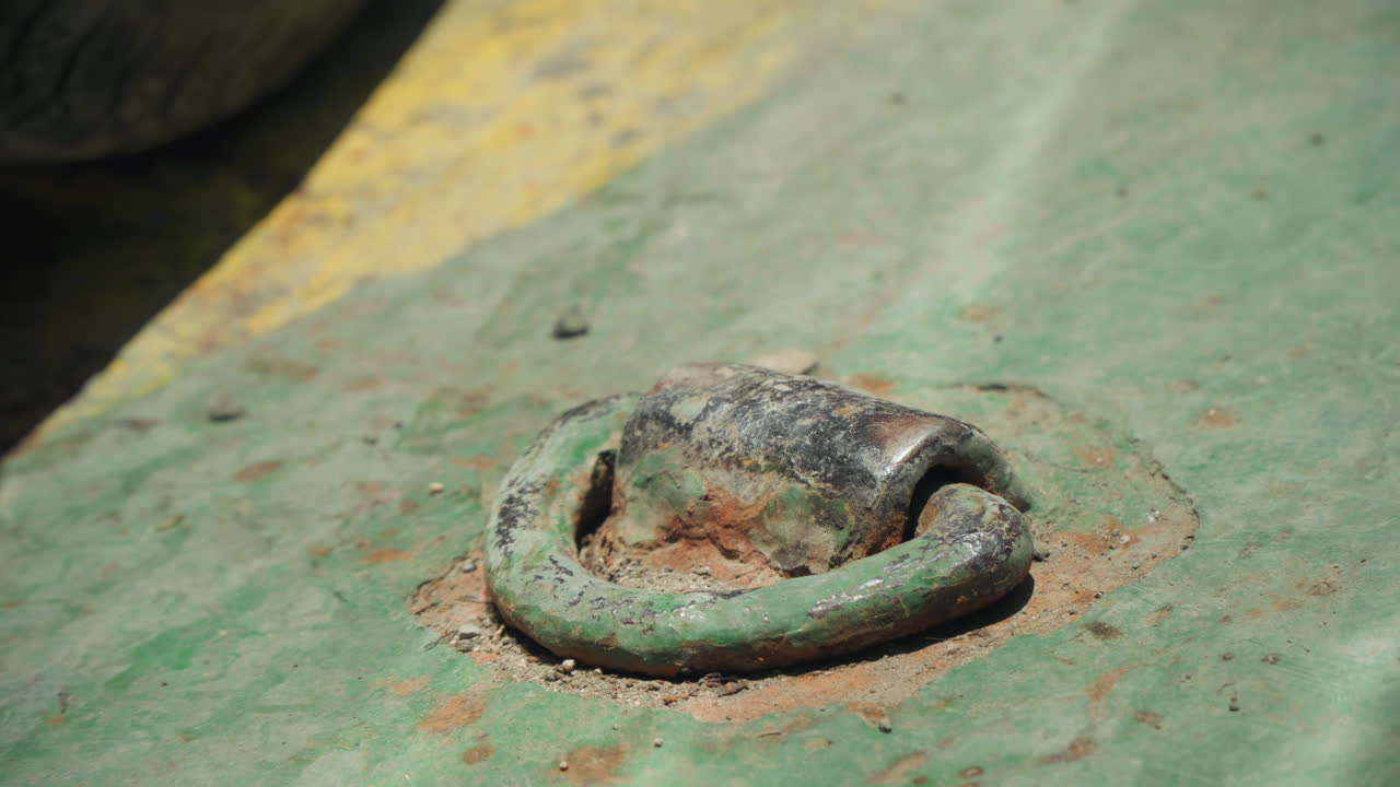 Primo piano di anello metallico arrugginito su piastra verniciata di verde, sabbia e ruggine a scaglie circondano il fissaggio ad anello a D, fissaggio per rimorchio industriale, striscia gialla sbiadita sul bordo, superficie granulosa e rovinata che trasmette incuria