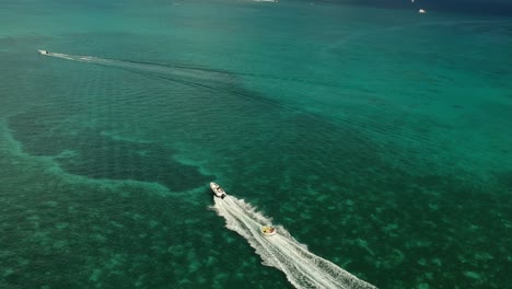 Aerial-shot-of-a-sportboat-pulling-a-big-bloat-with-people-on-it,-sailing-in-amazing-waters