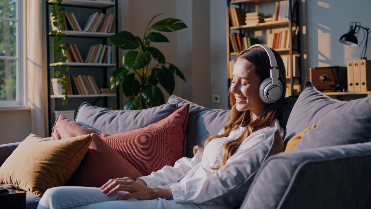 A relaxed woman enjoys music on headphones, sitting on a cozy sofa. The video captures a side angle