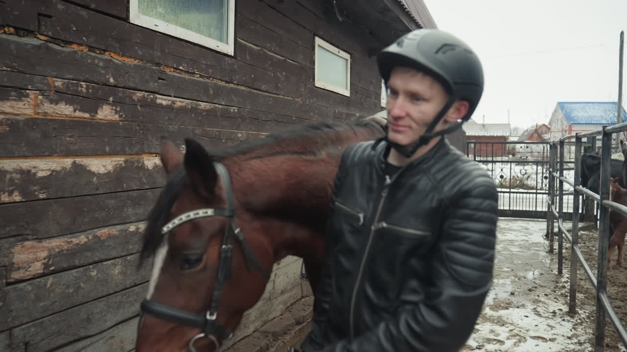 Uomo che addestra un cavallo in una stalla rustica. L'istruttore, con casco e giacca di pelle, conduce un cavallo marrone lungo un corridoio fangoso. Sottosella verde e briglia visibili in primo piano, neve invernale a terra, stalla di legno.