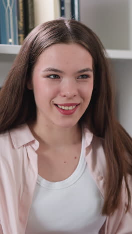 brunette woman smiles sitting by bookcase in library. pretty young lady sits on floor against bookshelf in store. cheerful student spends time in archive