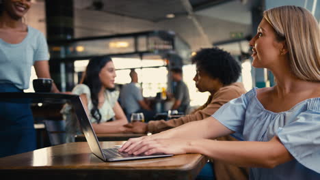 Mujer-Usando-Una-Computadora-Portátil-En-Una-Cafetería-Pagando-La-Factura-Con-Pago-Por-Teléfono-Móvil-Sin-Contacto