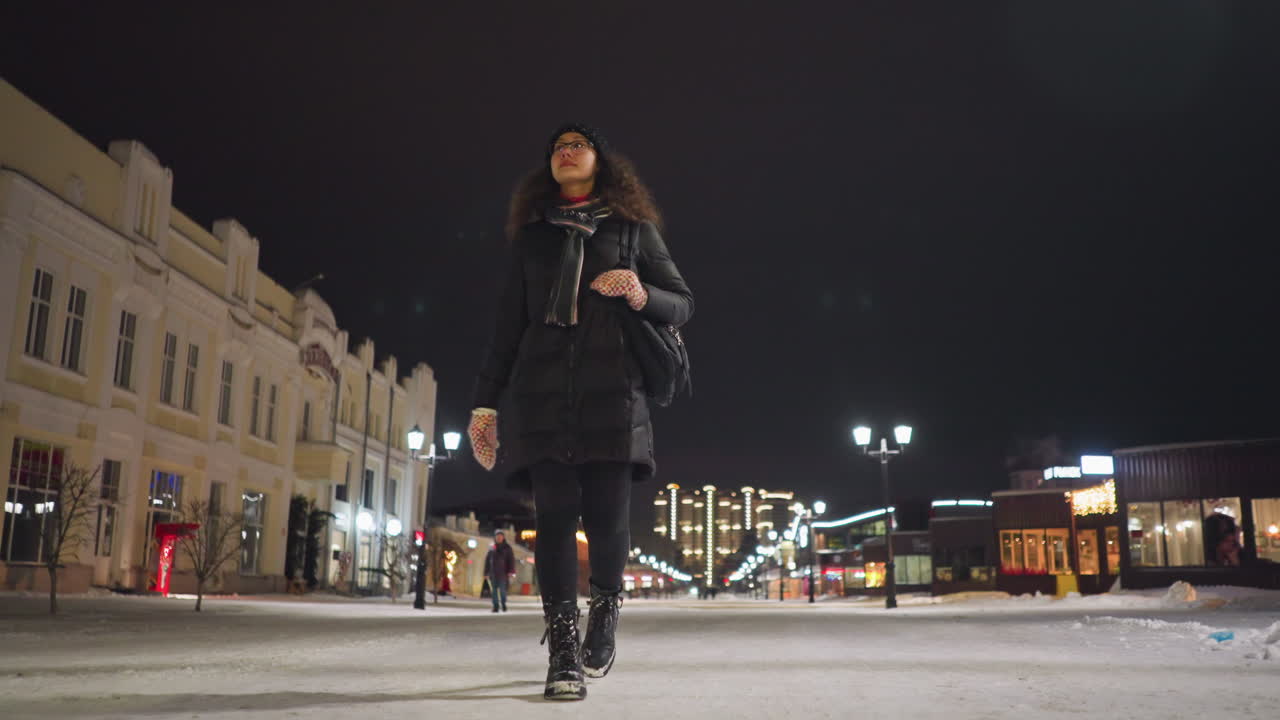 Woman in warm winter coat, scarf, and mittens walks alone on snowy city street at night, illuminated by glowing street lamps and festive lights, creating serene urban atmosphere with modern buildings