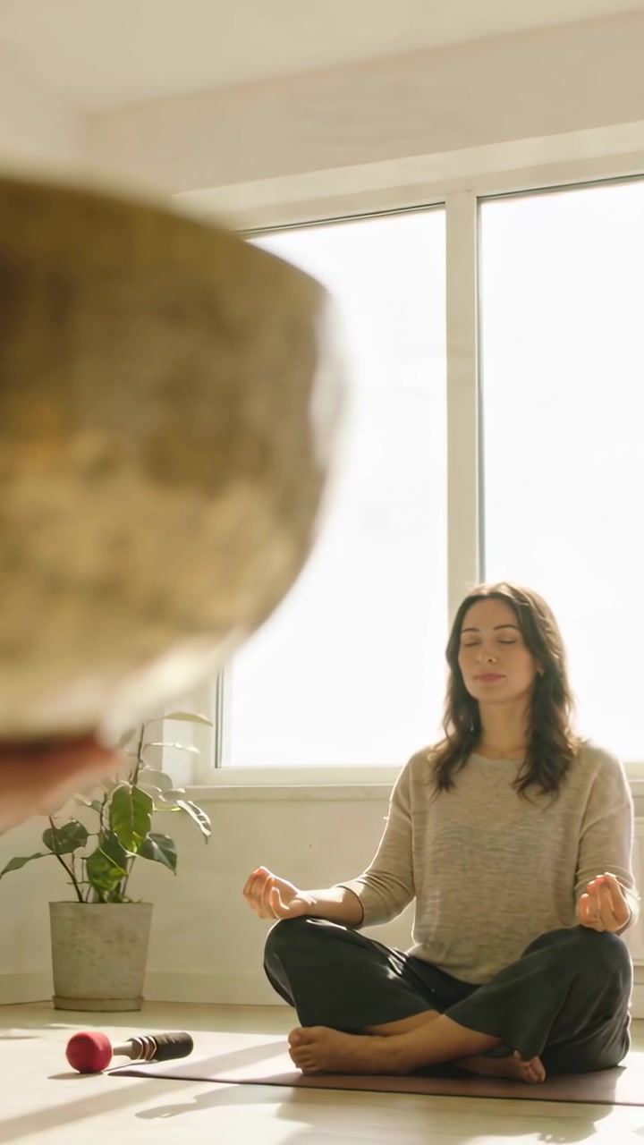 Video verticale: Una mano fuori campo colpisce il bordo di una ciotola tibetana su un tappetino, con una donna in top chiaro per la meditazione.