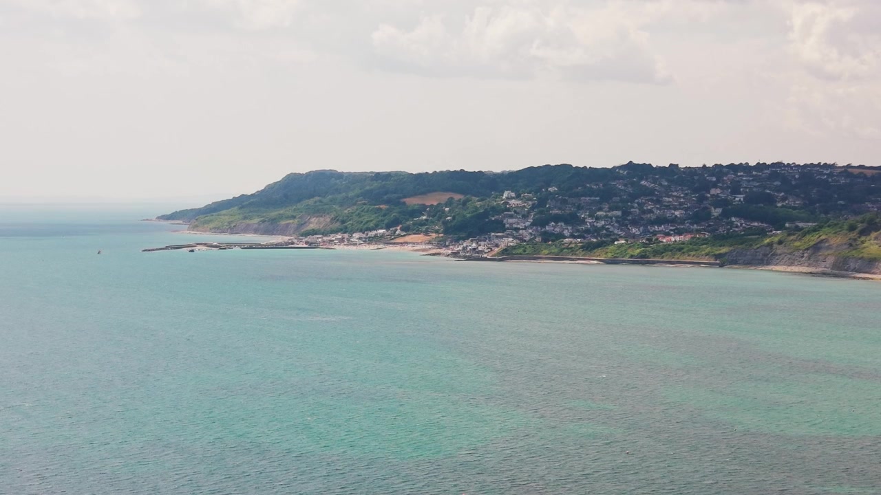 Riprese panoramiche con drone che guardano attraverso le acque calme della Baia di Lyme verso la città costiera di Lyme Regis nel Dorset, Inghilterra. L'ampia vista mostra case in collina, scogliere boscose e la costa.