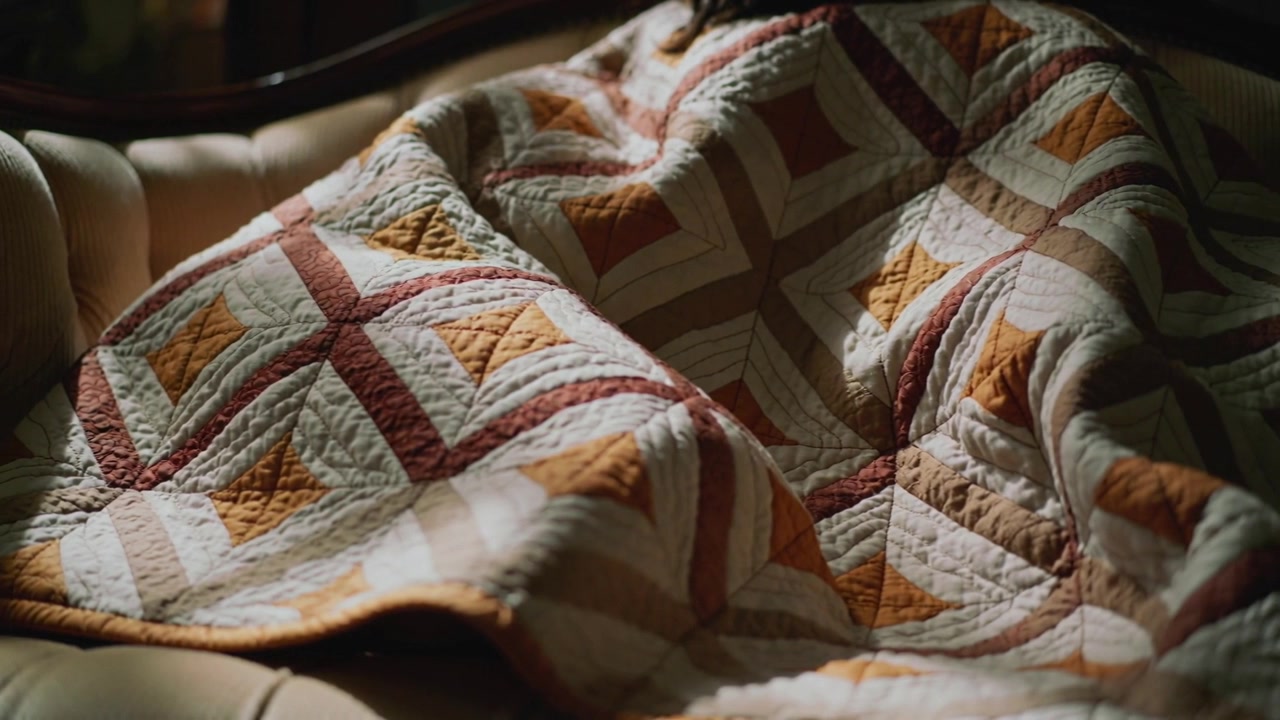 Shifting warm light making patchwork quilt settling folds on tufted armchair, showing stitches