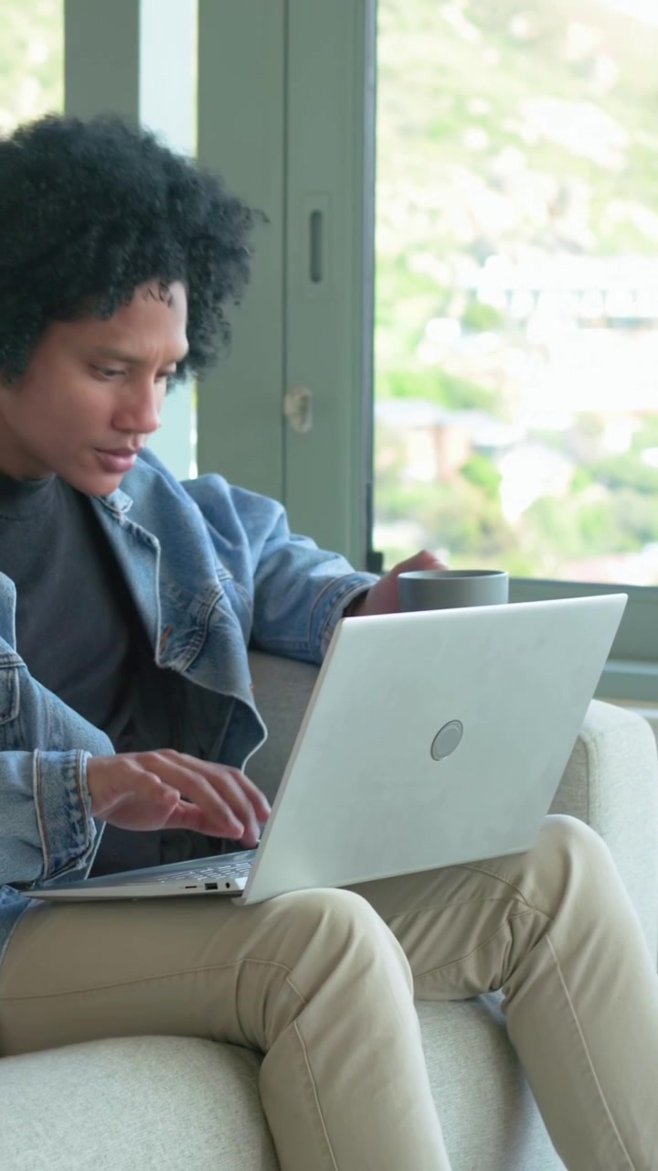 Vertical video: Leaning denim jacket man typing on laptop on couch by window, holding mug for work