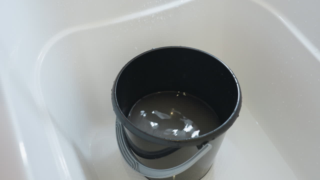 Close up of water streaming into black bucket placed in bathtub, filling container with splashing ripples, showing sanitation and household cleaning preparation