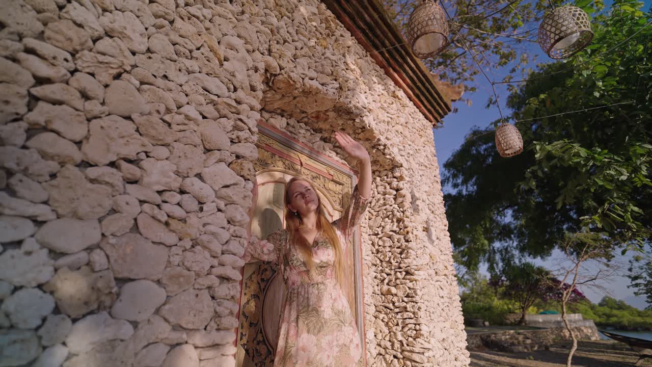 Eine Frau berührt sanft ihr Haar, während sie mit einer Hand ihre Augen vor der Sonne schützt, stehend vor einer Korallensteinmauer. Eine tropische Umgebung schafft eine warme, natürliche und Lifestyle-Atmosphäre.