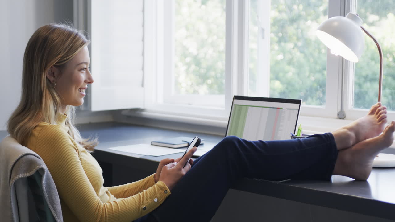 Portrait of happy biracial woman sitting at desk using smartphone and laptop at home, slow motion