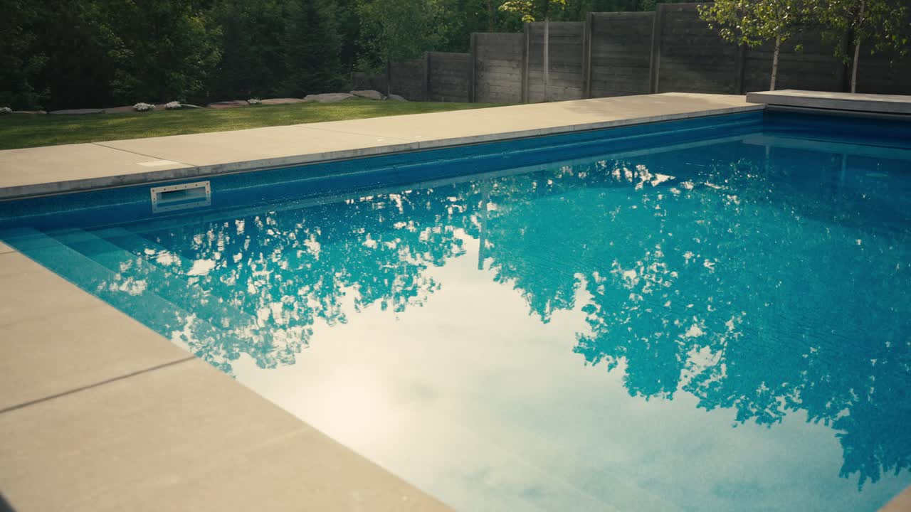 En swimmingpool i baghaven ved en luksusvilla med blåt vand og træerne og sollyset synligt i vandets refleksion
